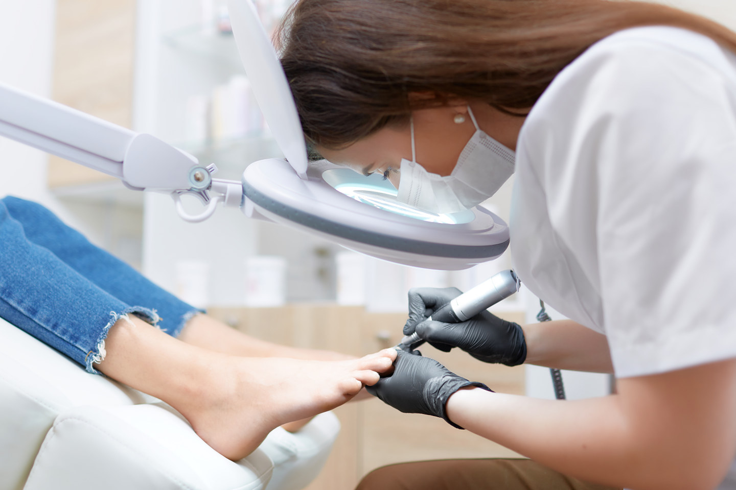 View of podiatrist doctor in white working at beauty salon, making polish procedure for foot with special equipment for blondie client. Young woman resting, caring about her health and foot.