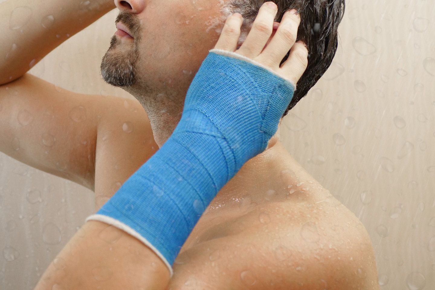 Handsome middle-aged man takes a shower with his hand wrapped in a plaster cast, washes his hair in the bathroom. Broken wrist in modern blue waterproof bandage